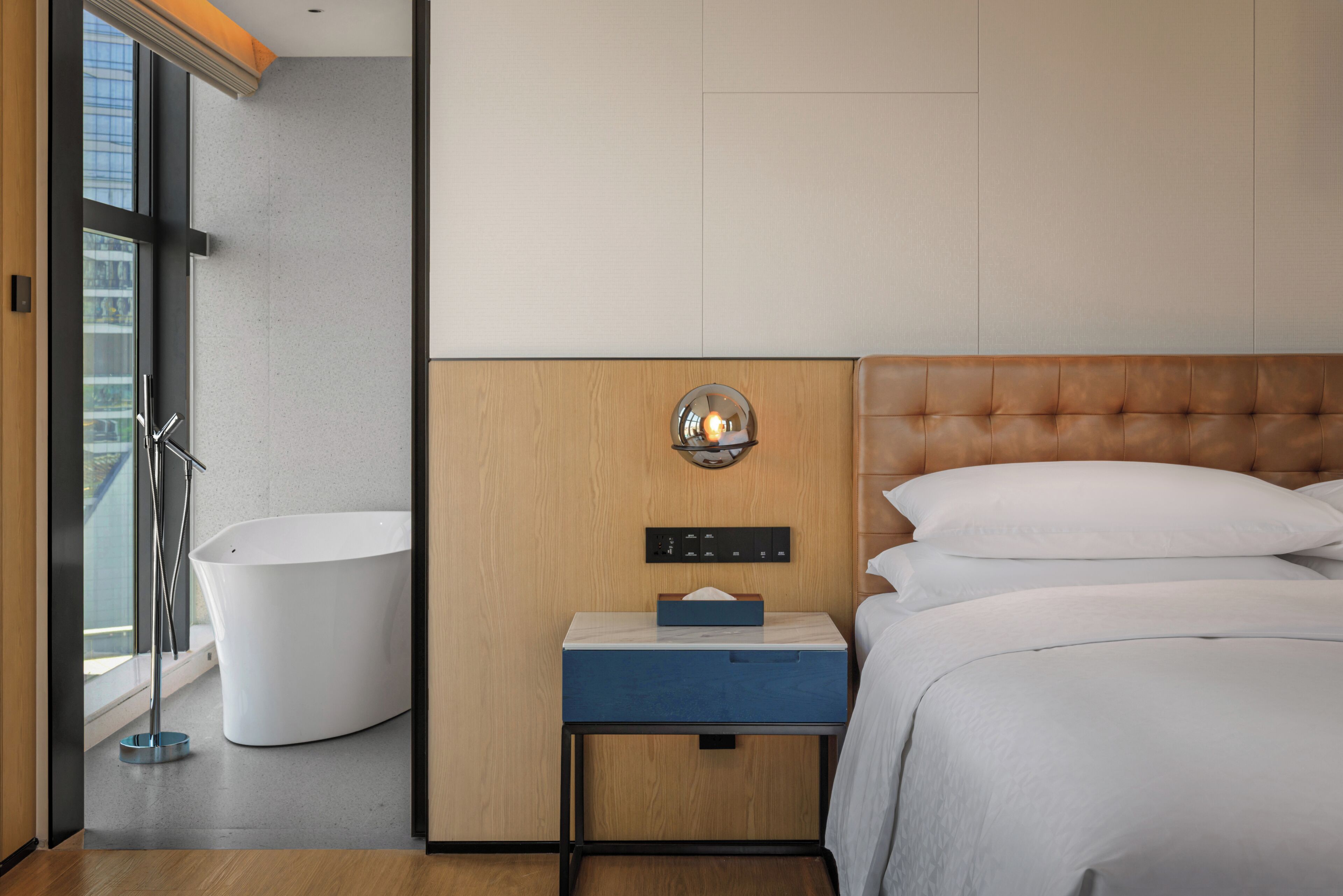 Een moderne hotelkamer met een badkuip, een nachtkastje met zakdoosjes en een groot bed met wit beddengoed.