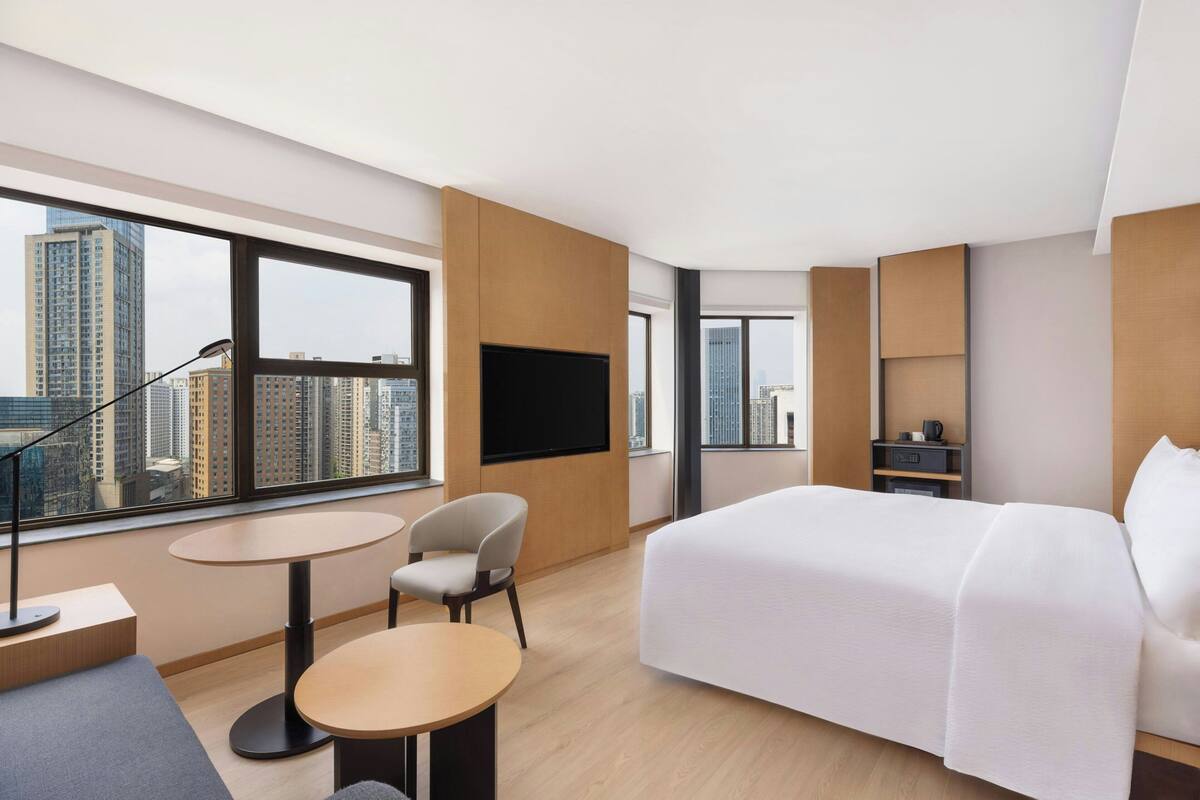A modern hotel room with a large bed, a desk, a chair, a TV, and a city view.