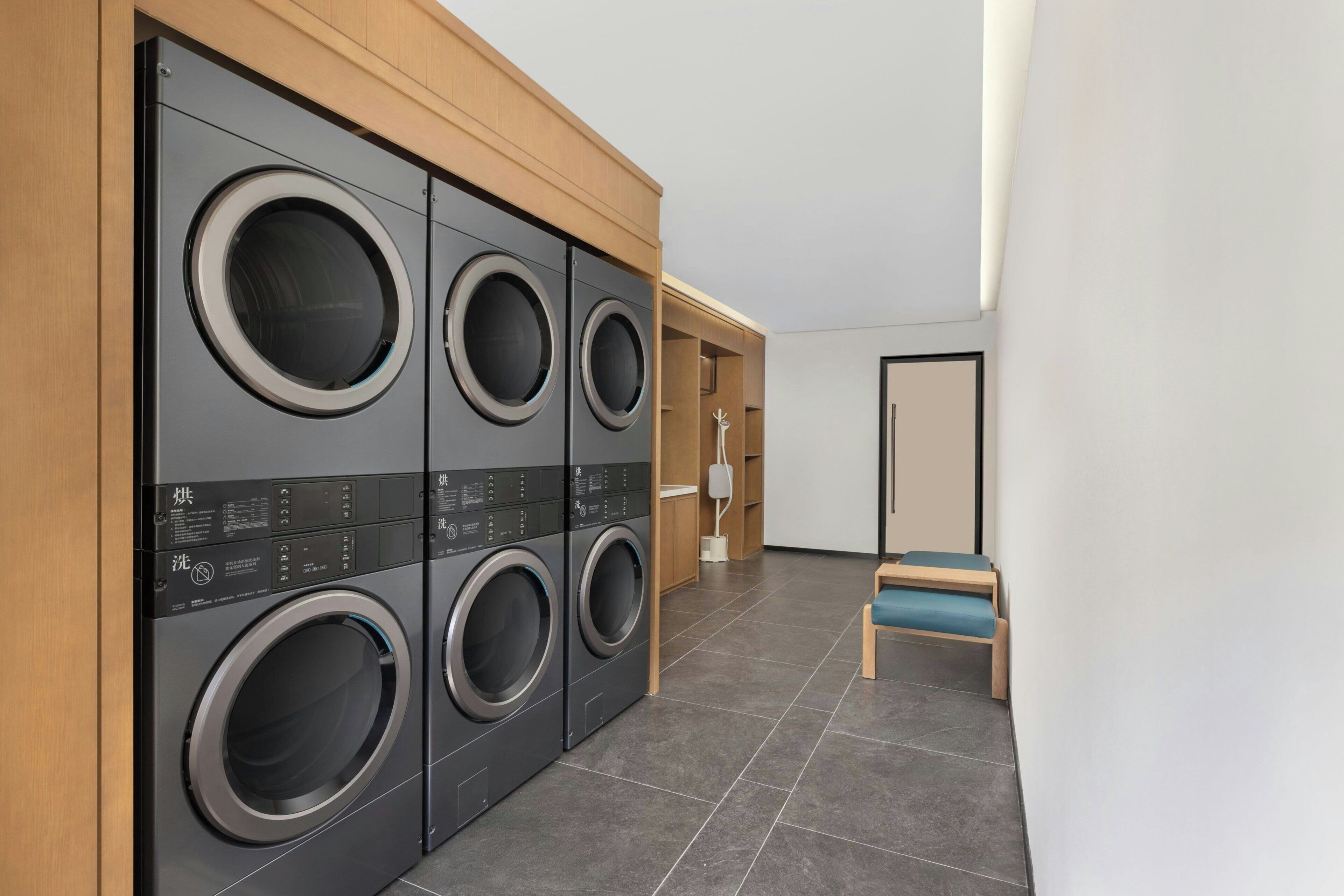 Un cuarto de lavado con cuatro lavadoras de carga frontal, un banco de madera y una puerta que da a otra habitación.