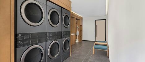 Un cuarto de lavado con cuatro lavadoras de carga frontal, un banco de madera y una puerta que da a otra habitación.
