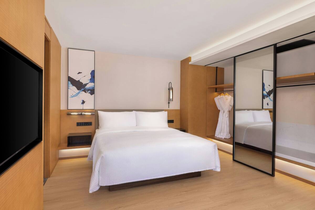 A modern hotel room with a large bed, a flat-screen TV, a bedside table, a robe, and a mirror.