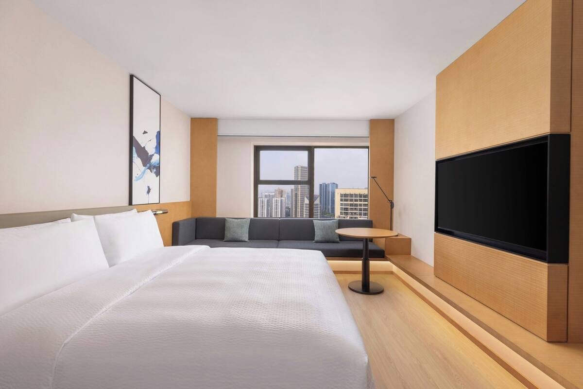 A hotel room with a large bed, a sofa, a TV, and a window with a city view.