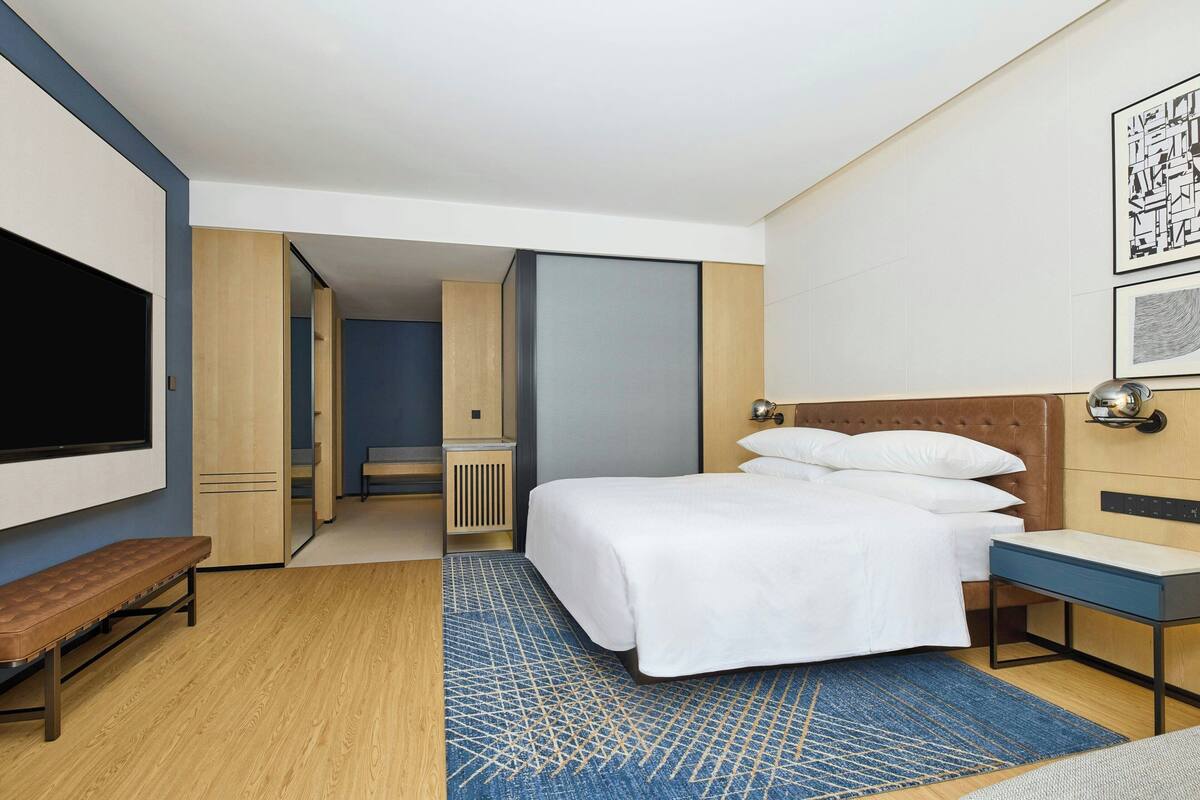 Quarto de hotel moderno com uma cama grande, um banquinho, uma mesa de trabalho, televisão de tela plana e um tapete azul estampado.