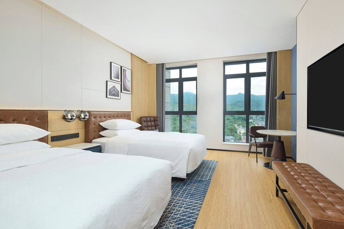 Quarto de hotel com duas camas, uma mesa, uma cadeira e uma janela grande com vista para as montanhas.
