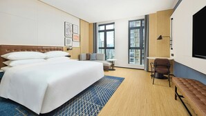 Una habitación de hotel con una cama grande, un escritorio, una silla, un banco y ventanas amplias.