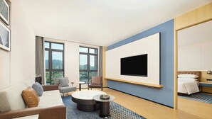 Habitación de hotel con una televisión grande, un sofá, una mesa de centro redonda, una silla, una cama y vistas al exterior.