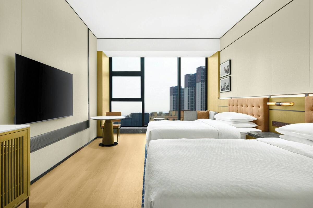 A hotel room with two beds, a flat-screen TV, a small table, and a window with a city view.