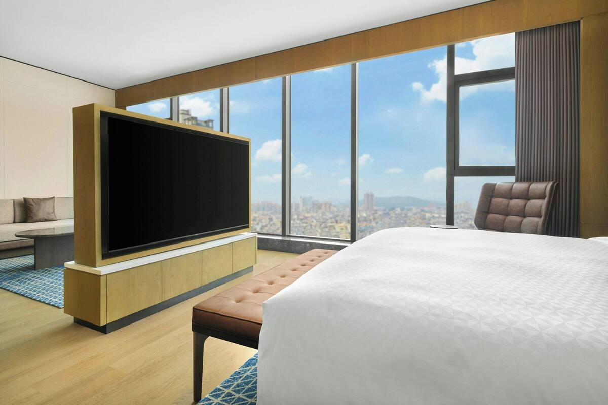 A modern hotel room with a large bed, a flat-screen TV, a seating area, and a city view.