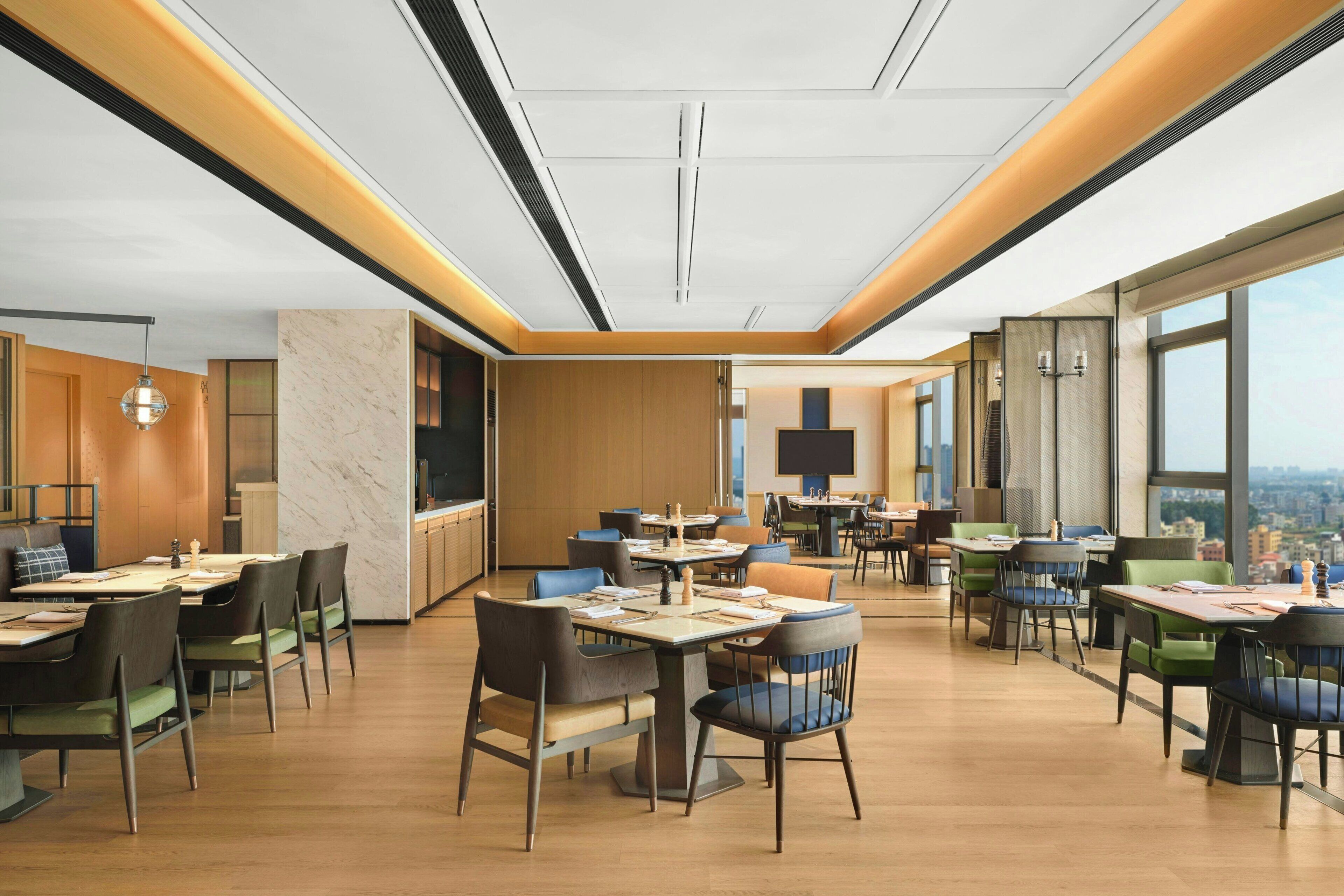 Un comedor moderno con varias mesas y sillas, una zona de cocina y ventanas amplias con vista a la ciudad.