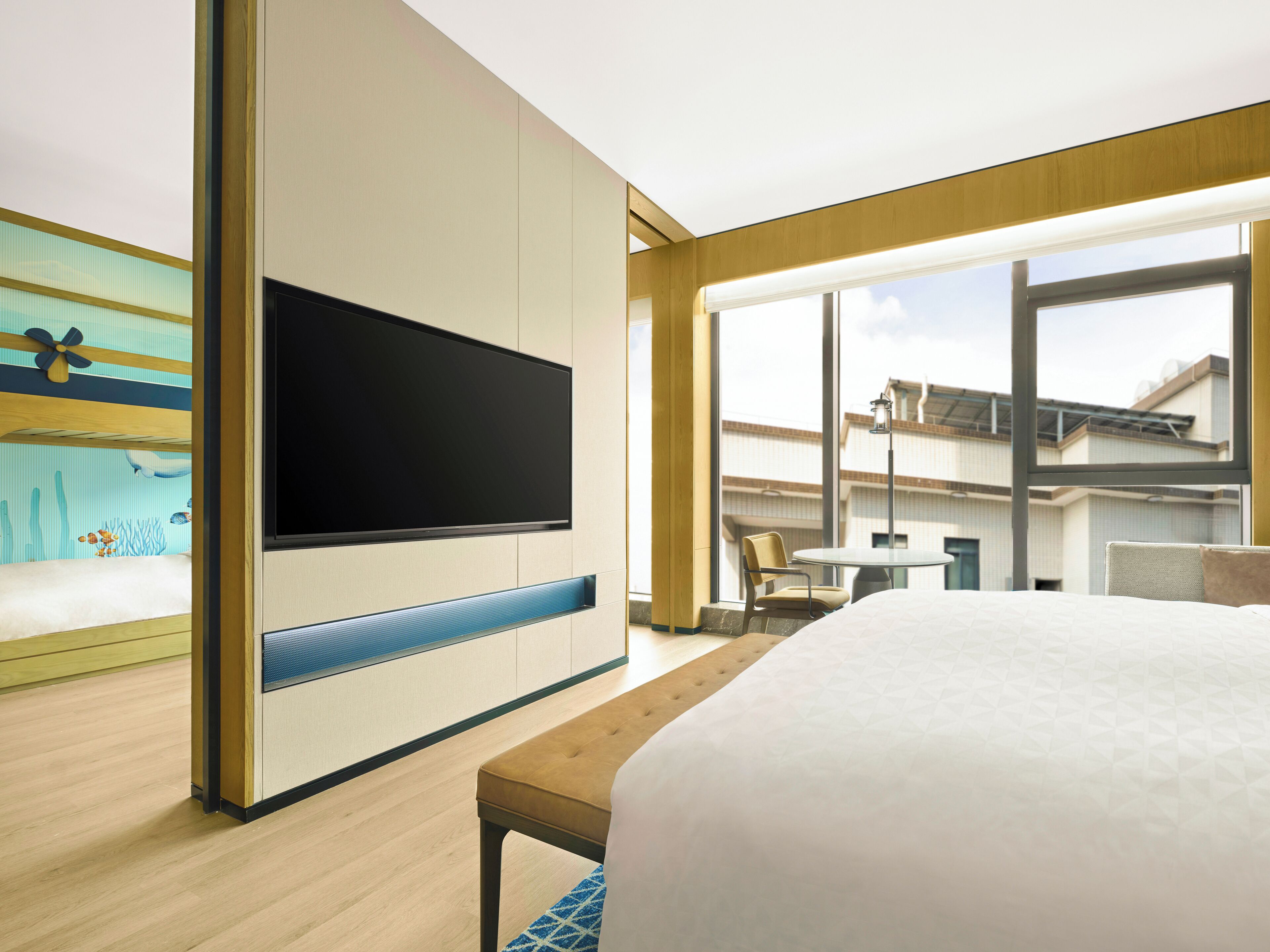 A modern hotel room with a large bed, a flat-screen TV mounted on the wall, a dining area with a table and chairs, and large windows offering a view of the building's exterior.
