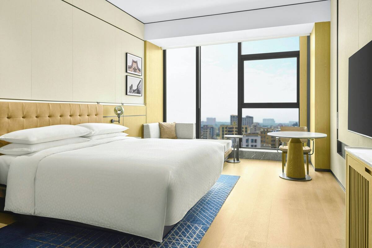 A hotel room with a large bed, a small table, a chair, and a city view through large windows.