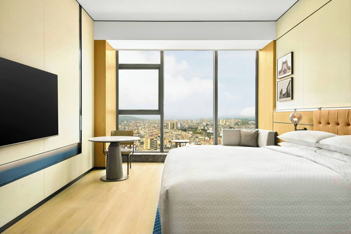 A hotel room with a large bed, a TV, a round table, a chair, and a city view through large windows.