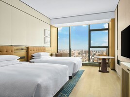 Habitación de hotel con dos camas, un ventanal con vistas a la ciudad, una mesita y un televisor de pantalla plana.