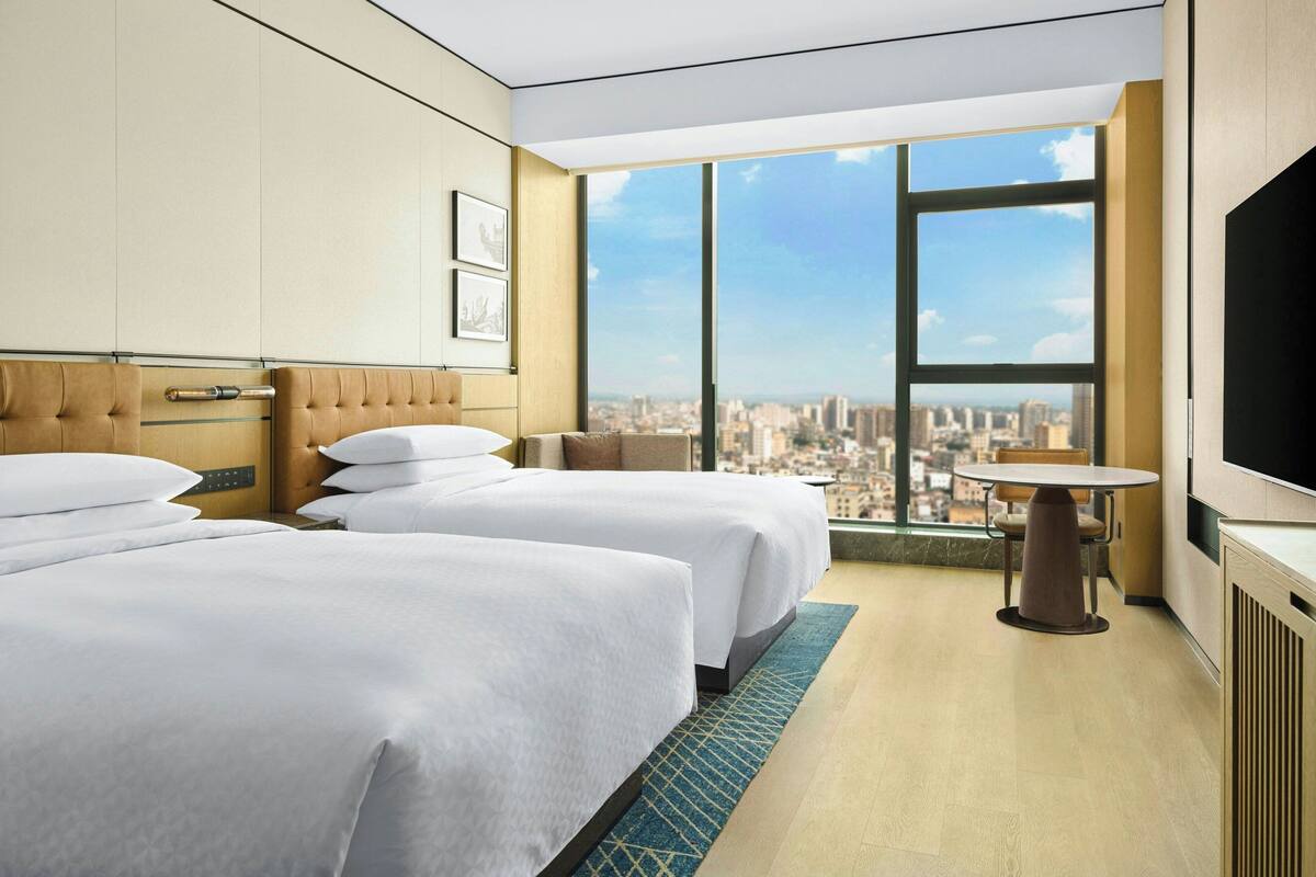 A hotel room with two beds, a large window with a city view, a dining table, and a flat-screen TV.