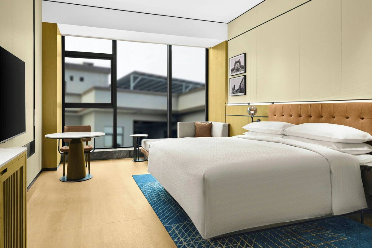 A hotel room with a large bed, a small table, a chair, a TV, and a window with a view of buildings.
