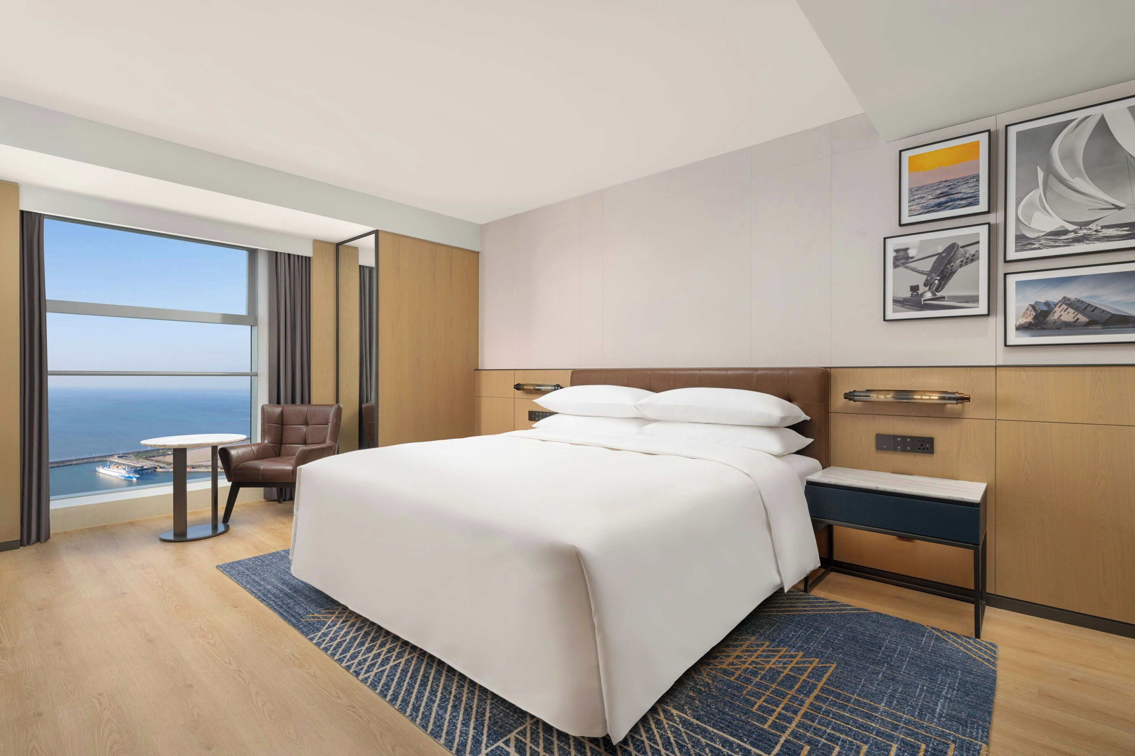 A hotel room with a large bed, a desk, a chair, a view of the ocean, and artwork on the wall.