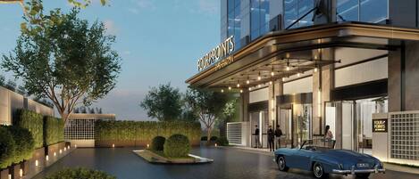 A modern hotel entrance with a car parked in front, a well-lit facade, and a landscaped area with trees and shrubs.