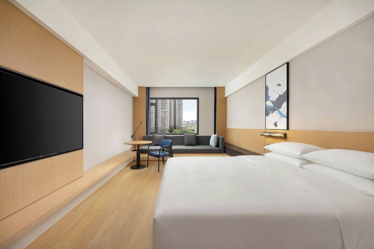 A modern hotel room with a large bed, a desk, a chair, a flat-screen TV, and a window with a city view.