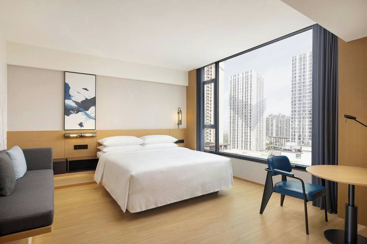 A hotel room with a bed, a desk, a chair, a large window with a city view, and a wall-mounted artwork.