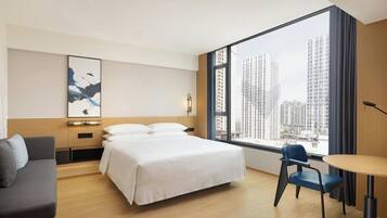 A hotel room with a bed, a desk, a chair, a large window with a city view, and a wall-mounted artwork.