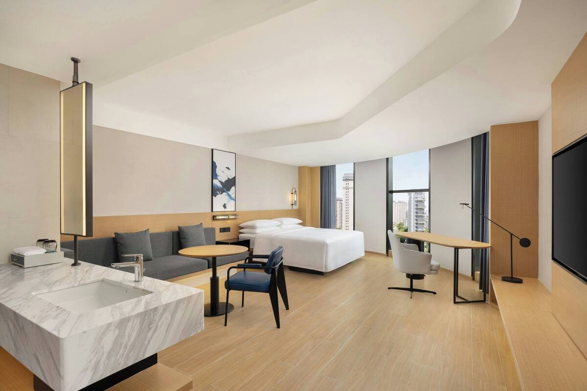 A modern hotel room with a marble sink, a sofa, a bed, a desk, and a large window.