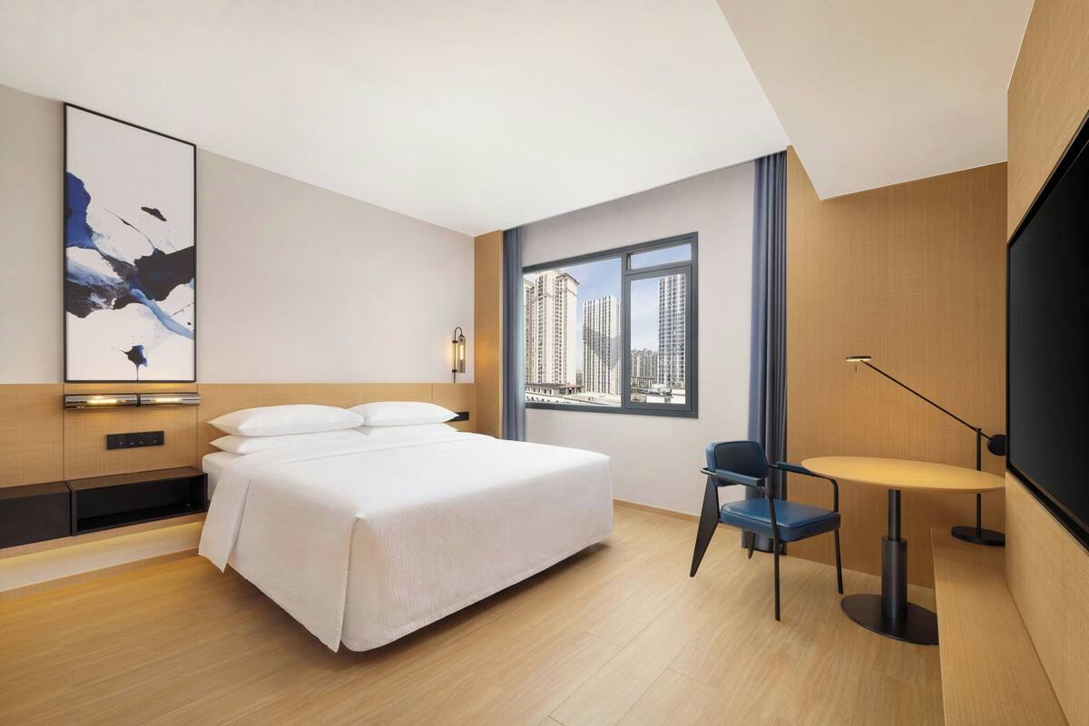 A modern hotel room with a large bed, a desk with a chair, a large window with a city view, and a wall-mounted artwork.
