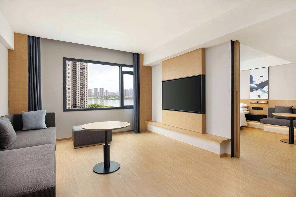 A modern hotel room with a large window, a flat-screen TV mounted on the wall, a round dining table, and a sofa.