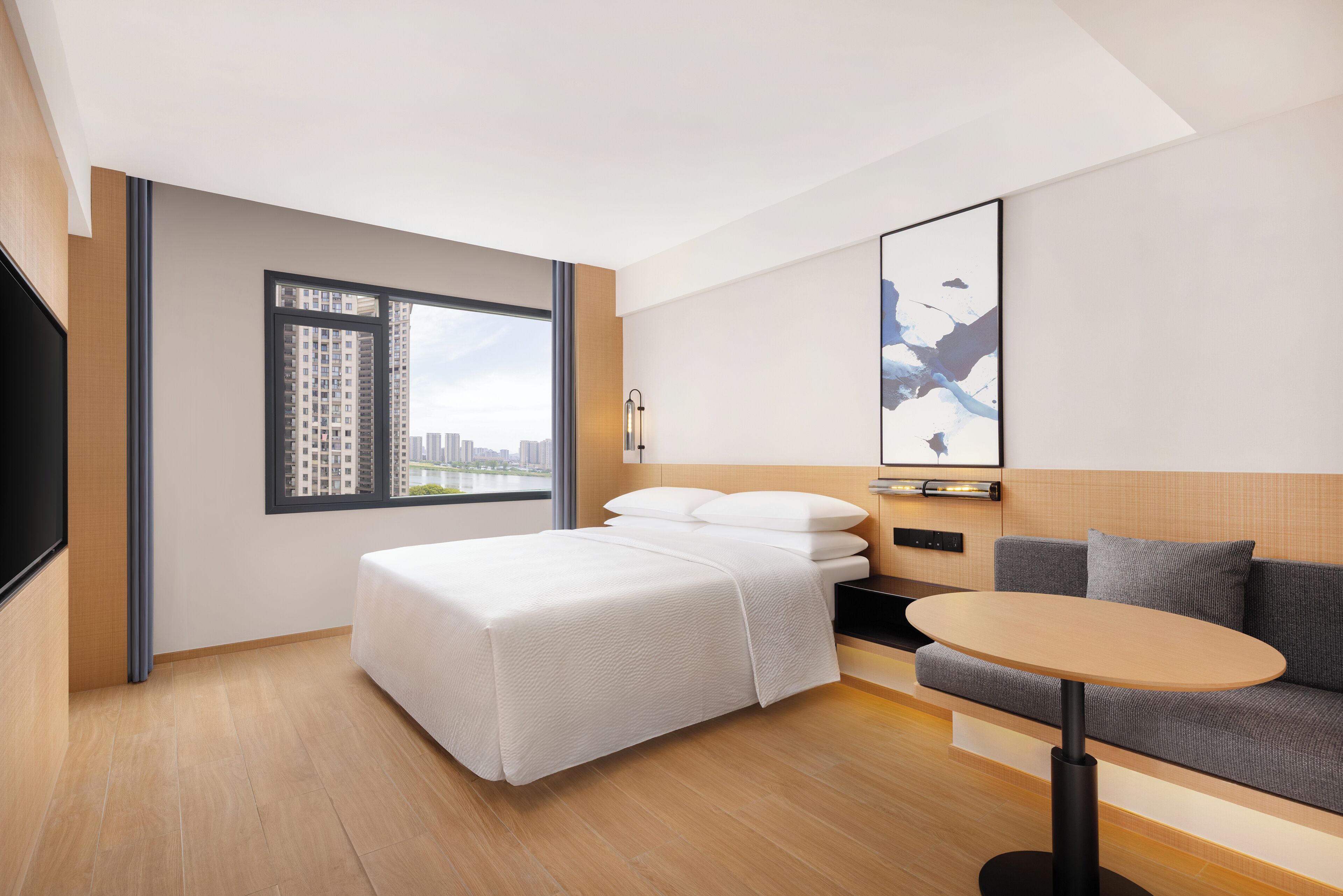 Una habitación de hotel moderna con cama, sofá, una mesita, un ventanal con vistas a la ciudad y una obra de arte enmarcada en la pared.