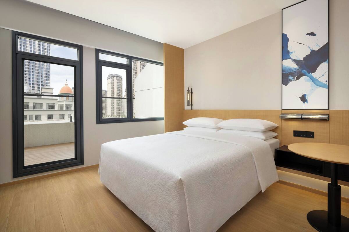 A hotel room with a bed, a small table, a wall-mounted light, and a large window with a city view.