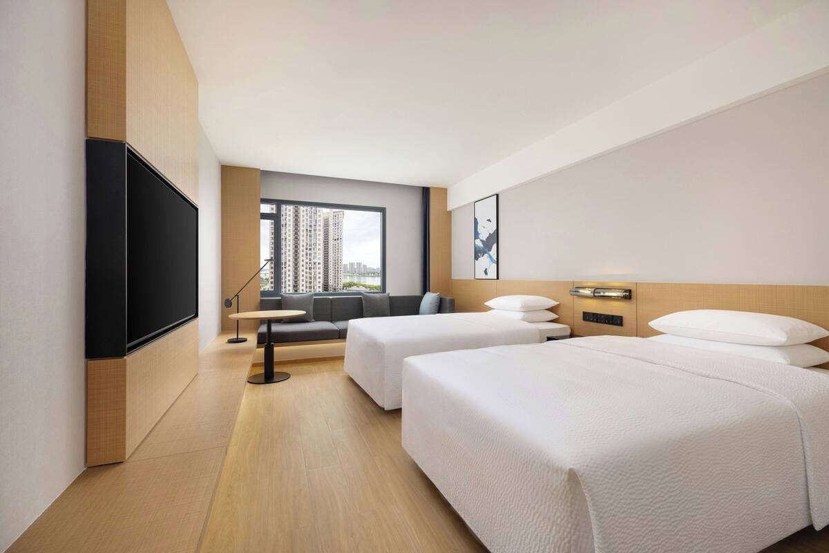 A hotel room with two beds, a sofa, a TV, and a window with a city view.