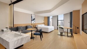 A modern hotel room with a marble bathroom, a sofa, a round dining table, and a large window offering a city view.