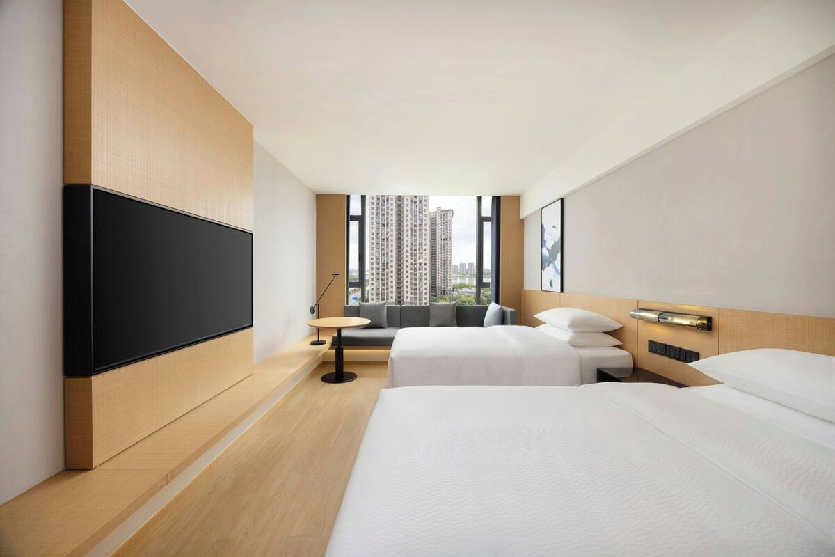 A hotel room with two beds, a TV, a small table, and a view of tall buildings.