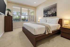 Une chambre à coucher comprenant un lit, des lampes de chevet, une table de nuit, une fenêtre avec des stores, un téléviseur fixé au mur et un tableau encadré.