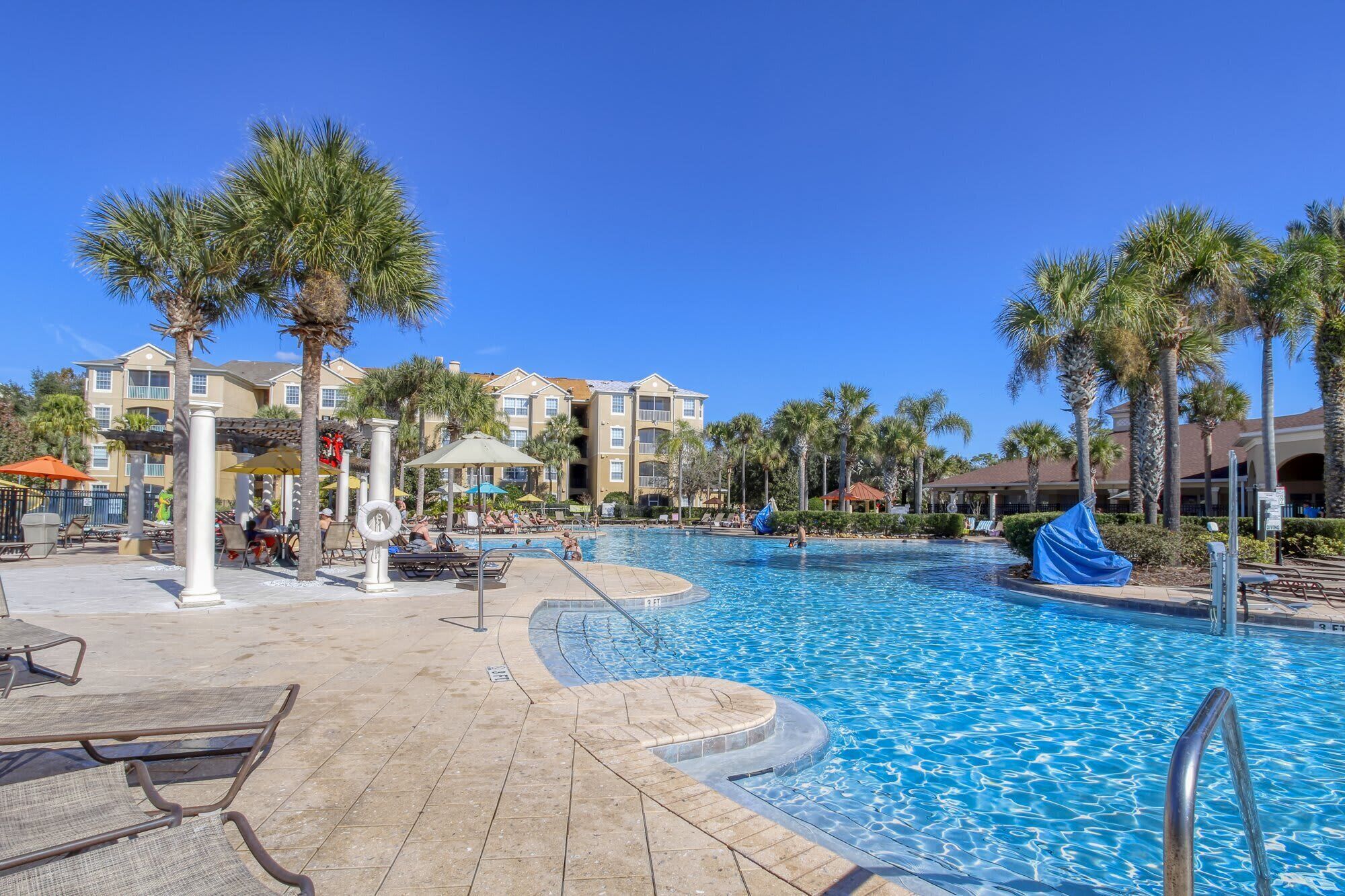 A resort pool area with lounge chairs, umbrellas, and palm trees.
