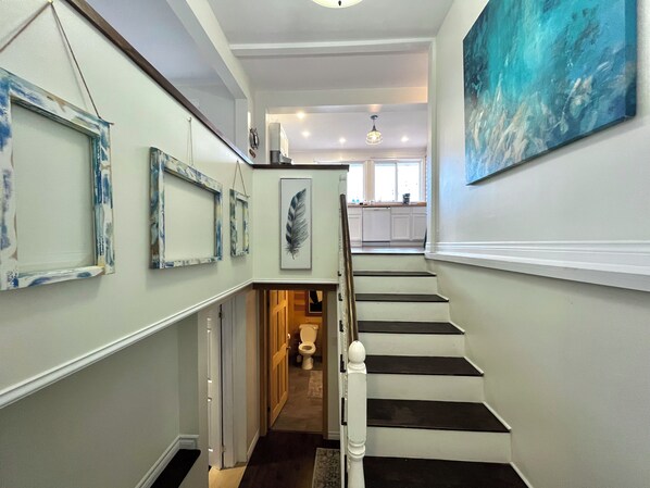 A staircase leading to a kitchen with white cabinets and a large painting on the wall. - Stay on the Coast | Steps to EC Trails, Private Torbay Retreat | Family Friendly (Torbay)