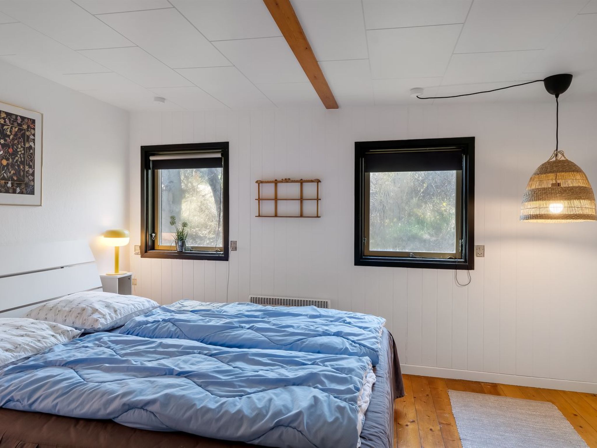 Un dormitorio con cama, dos ventanas, una lámpara fijada a la pared y un radiador.