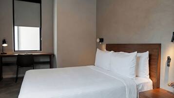 A neatly made bed with white linens, a wooden headboard, a desk with a chair, and a wall-mounted light fixture.