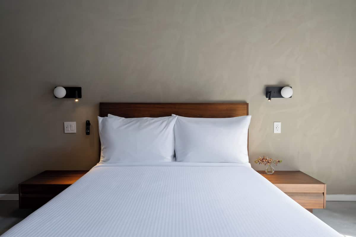 Una cama bien tendida con sábanas blancas, un cabecero de madera y dos mesitas de noche con lámparas de pared.