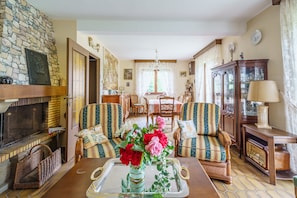 Uma sala de estar aconchegante com lareira de pedra, sofás listrados, uma área de jantar ao fundo e um vaso decorativo com flores sobre uma bandeja.