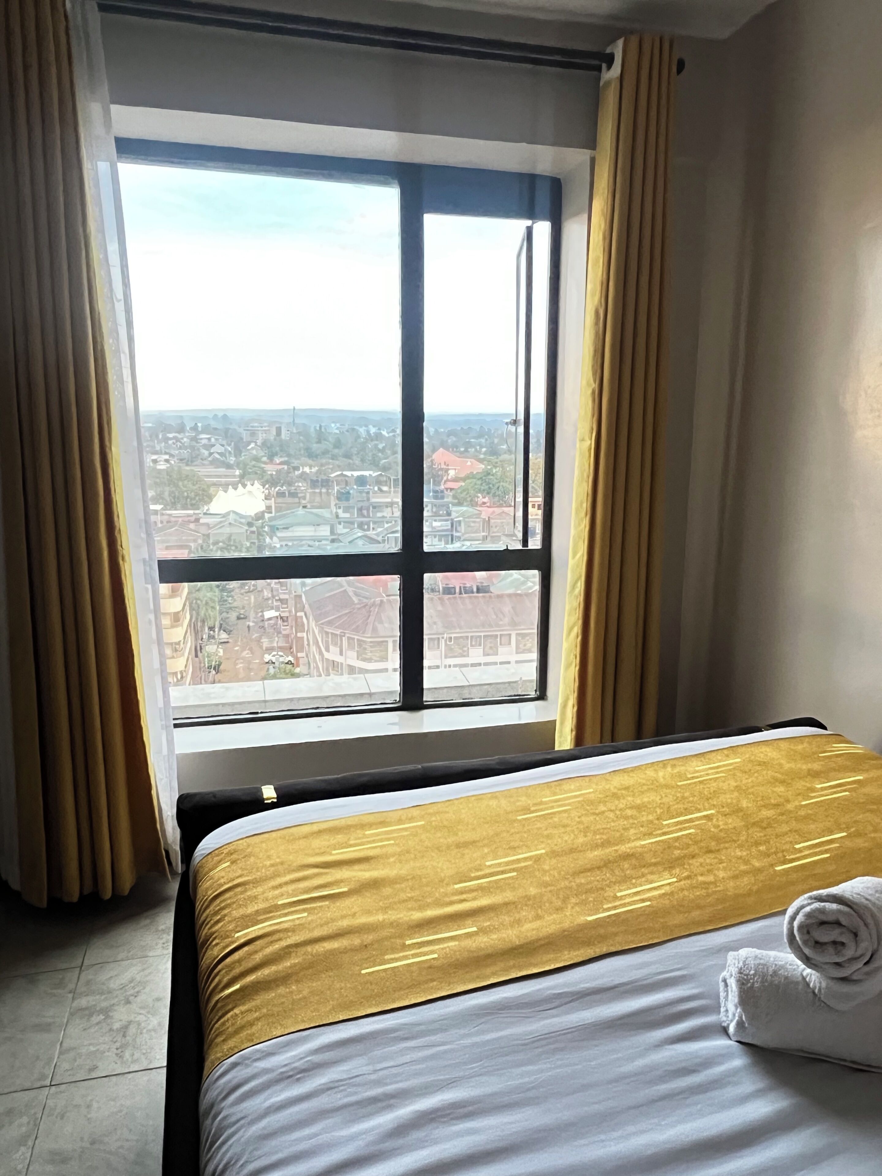 Une chambre d’hôtel avec une grande fenêtre, un lit recouvert d’une courtepointe aux motifs dorés et blancs, et une serviette enroulée posée sur le lit.