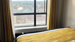Quarto de hotel com uma grande janela, uma cama com colcha dourada e branca, e uma toalha enrolada sobre a cama.