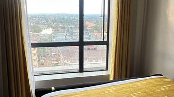 Une chambre d’hôtel avec une grande fenêtre, un lit recouvert d’une courtepointe aux motifs dorés et blancs, et une serviette enroulée posée sur le lit.