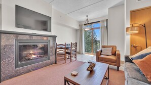 A living room with a fireplace, a television, a dining area, and a balcony with a view. - Fairway Hideaway - Golf Villa | HotTub | Waterpark (Wisconsin Dells)