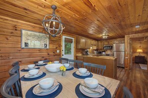A wooden cabin interior with a dining table set for six, kitchen area, and a large chandelier. - Escape to a warm and inviting Smoky Mountain cabin just minutes from downtown Gatlinburg. Enjoy peaceful mountain views, unwind in the hot tub, and gather with family or friends in a spacious, comfortable setting designed for relaxation and fun. (Gatlinburg)