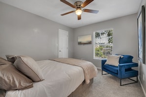 A bedroom with a bed, a blue chair, a ceiling fan, a window, and a door.