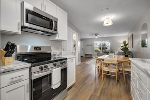 A modern kitchen with white cabinetry, a stainless steel stove, a microwave, and a dining area with wooden chairs.