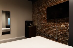 A room with a brick wall, a flat-screen TV mounted on it, a bed, and a small wooden cabinet. - Sosuite | Queen Room w Desk, Shared Roof Decks (Philadelphia)