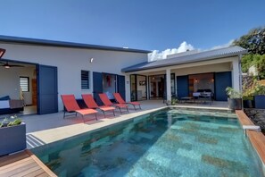A modern house with a swimming pool, red lounge chairs, and a covered patio area.