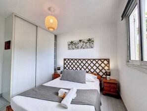 A neatly made bed with a headboard, bedside tables, a wall-mounted artwork, and a window with blinds. - Perle d'été - Balcon - Wifi (Saint-Paul)