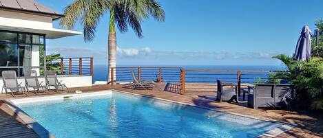 A pool area with lounge chairs, a palm tree, and a view of the ocean.